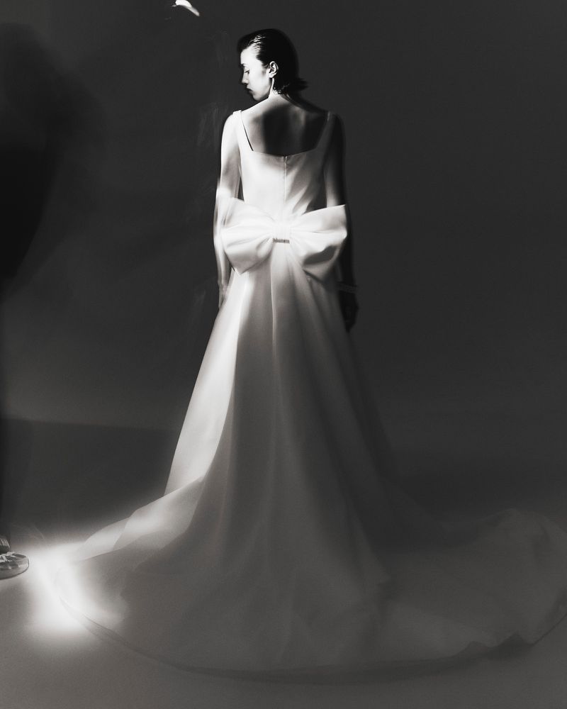 Woman in a white wedding gown with a bow stands in a photo studio with soft lighting, captured in a medium shot with a dark background.