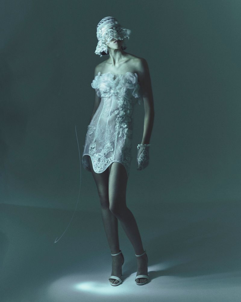 Woman in strapless white lace dress, matching hat and gloves, and high heels stands in a photo studio with cool lighting.