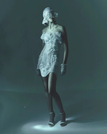Woman in strapless white lace dress, matching hat and gloves, and high heels stands in a photo studio with cool lighting.