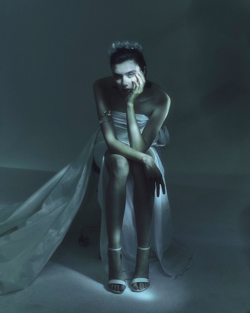 A woman in a white dress sits in a dimly lit studio with a crown, her face resting on her hand in a contemplative pose.
