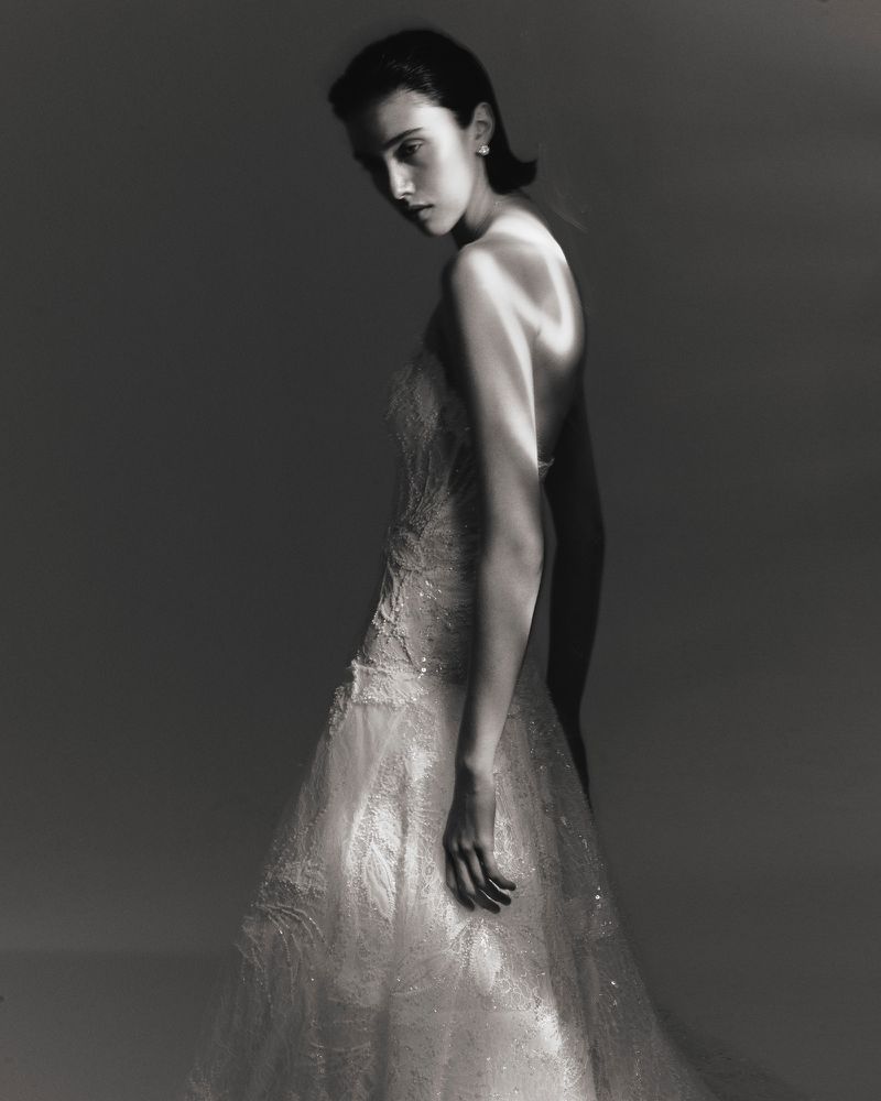 Model Sandra Formanek wearing a strapless embellished lace gown in dramatic studio lighting against a dark grey backdrop.