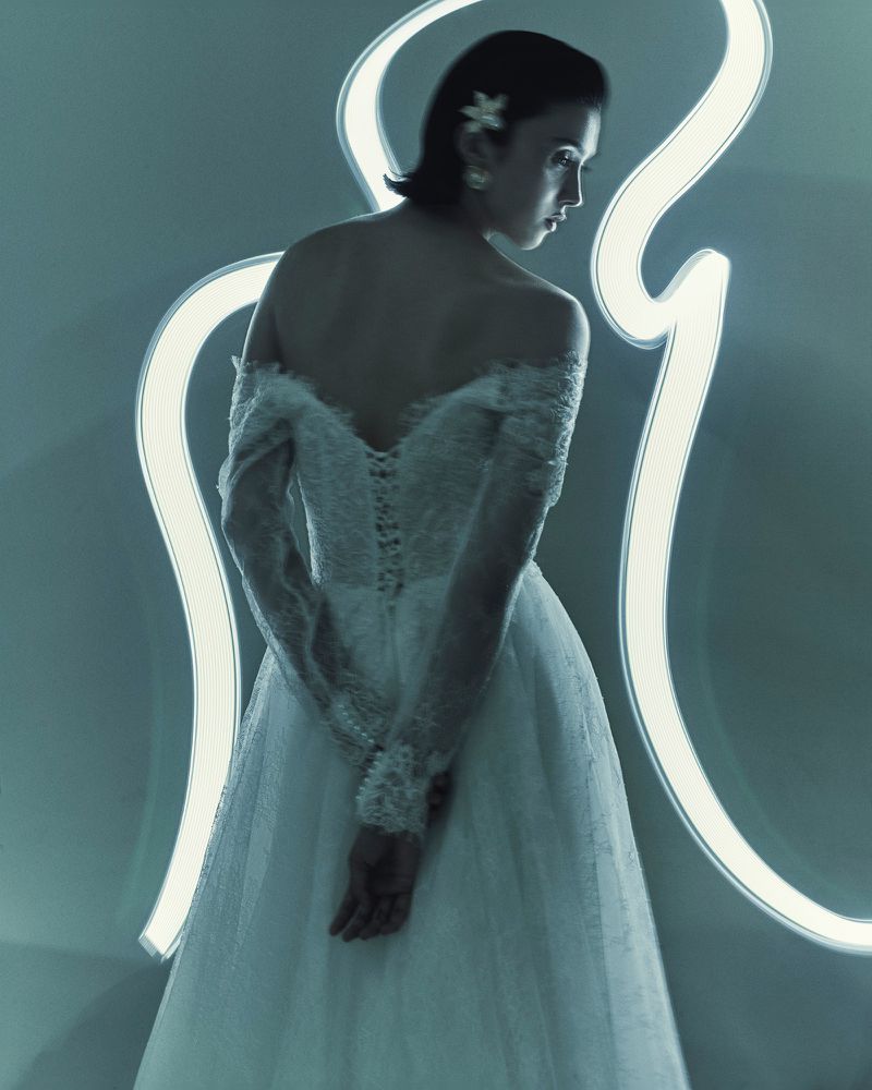 Woman in white off-the-shoulder gown with lace details, flower in hair, and neon light in background, shot in a photo studio.