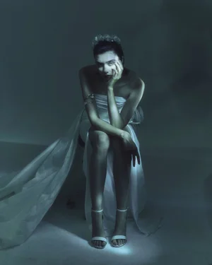 Model Sandra Formanek seated in a strapless white gown and strappy heels against a moody cool-toned studio background.