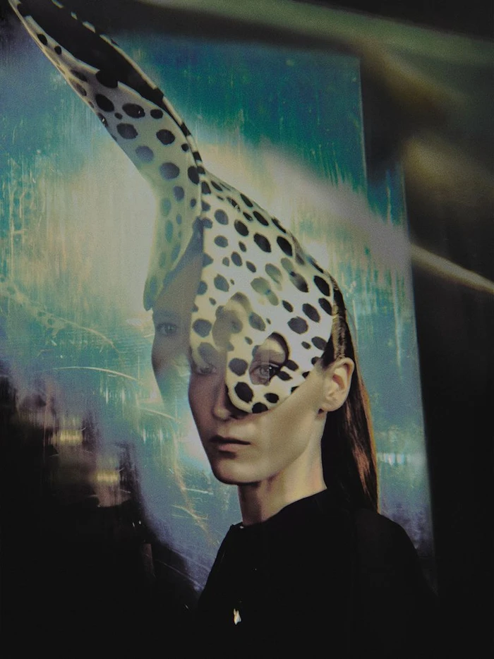 Model wearing a black top and a sculptural spotted headpiece against an abstract teal and blue background