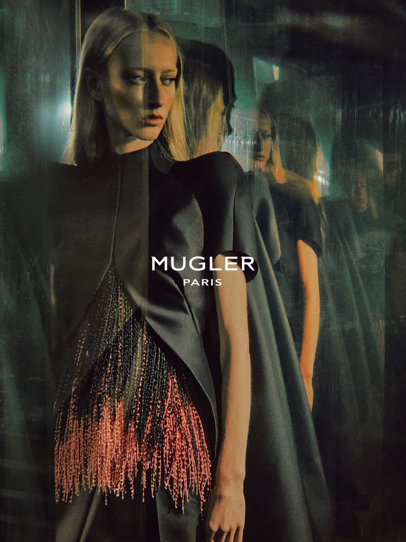 Model wearing dark structured attire with fringing, reflected multiple times in glass against a dark background