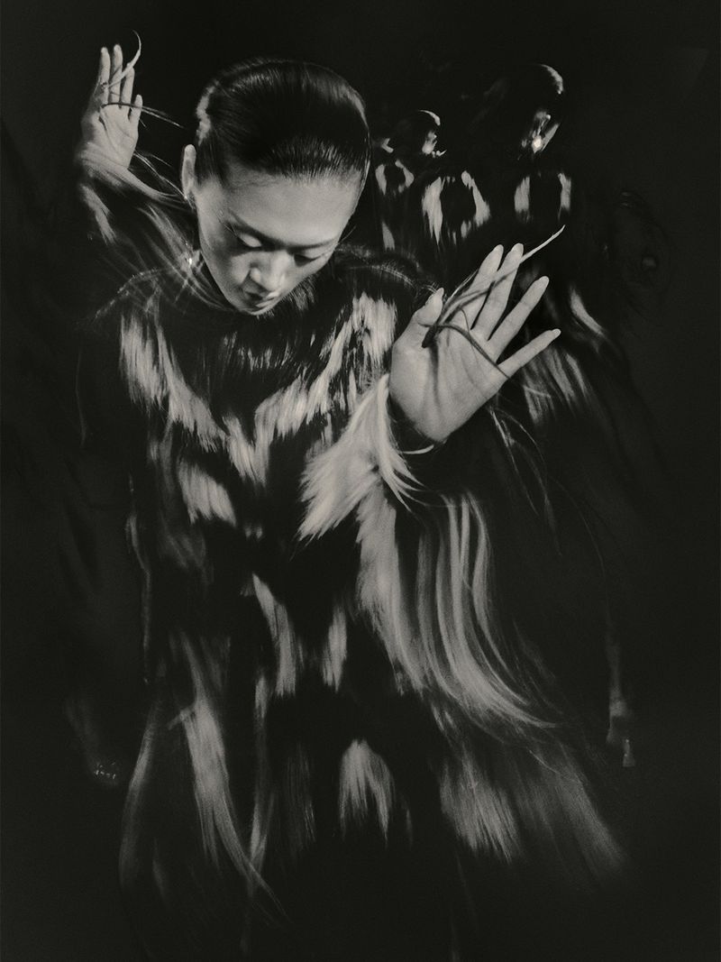 Model with dark slicked hair wearing a patterned garment with hands raised in motion against a dark background
