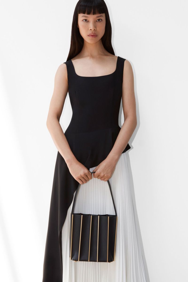 Asian woman posing in a black top and white pleated skirt, holding a black and gold handbag in a white studio setting.