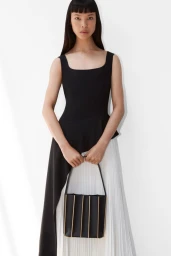 Model in black sleeveless top and white pleated skirt holding a black handbag against a white background