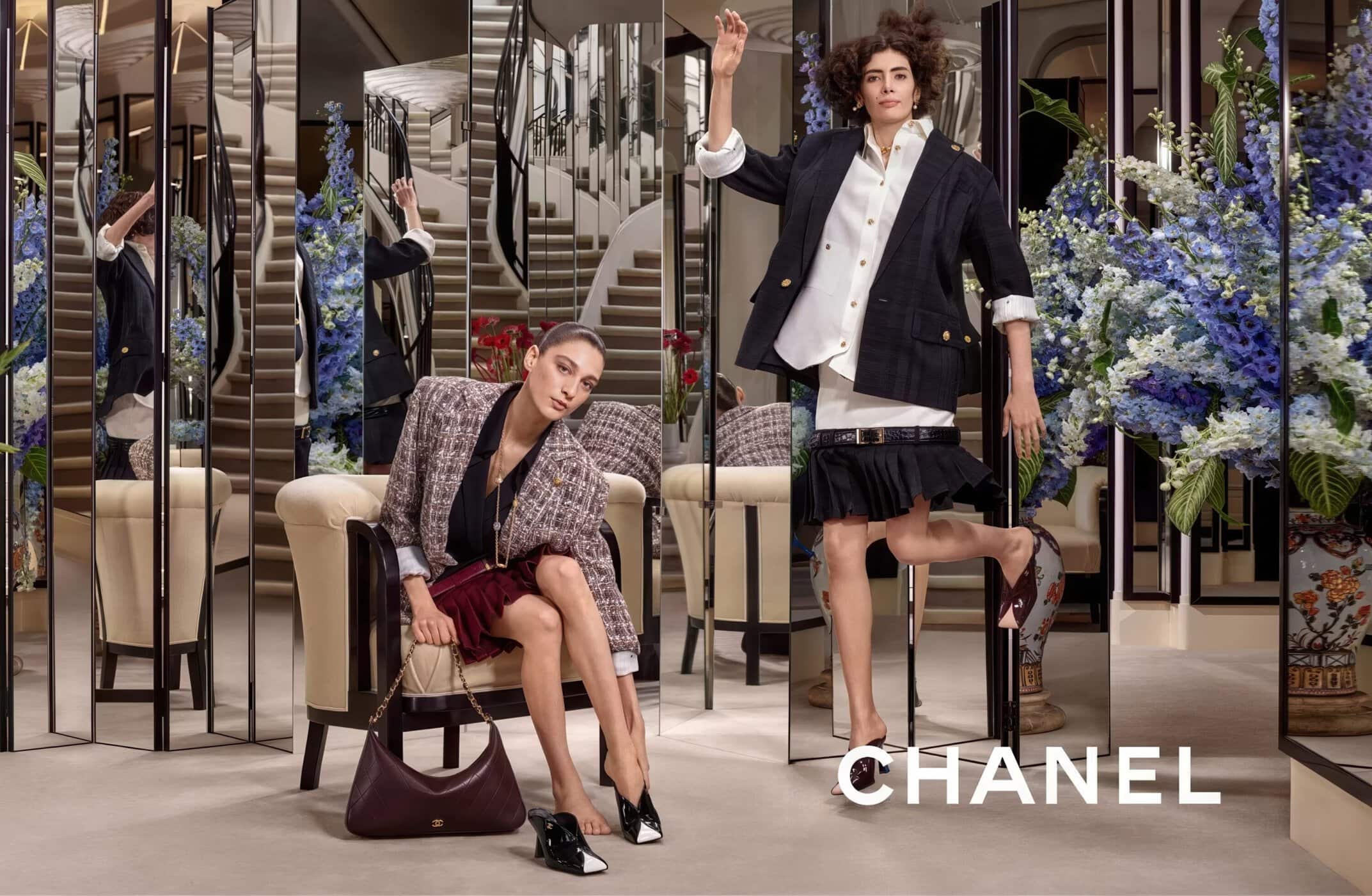 Two models posing in an interior space with mirrored panels, armchairs, and large floral arrangements for Chanel