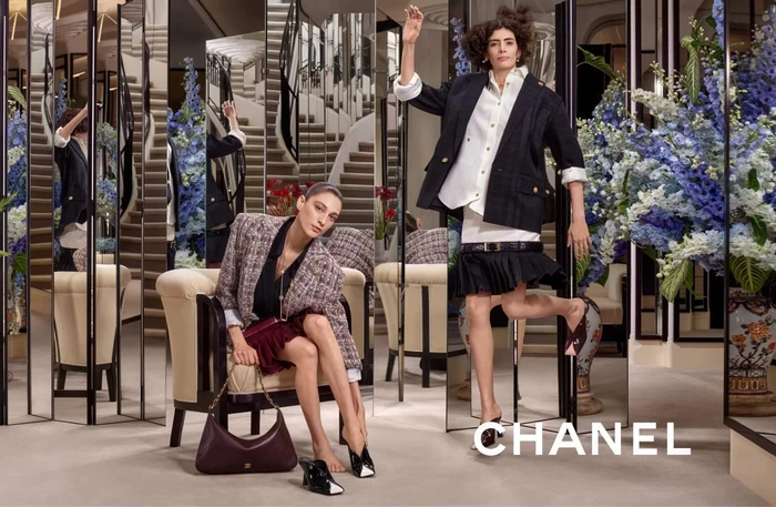 Two models posing in an interior space with mirrored panels, armchairs, and large floral arrangements for Chanel