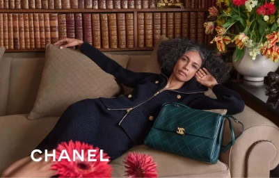 Model reclining on a sofa next to a green handbag in front of a bookshelf with red and yellow flowers in the foreground.