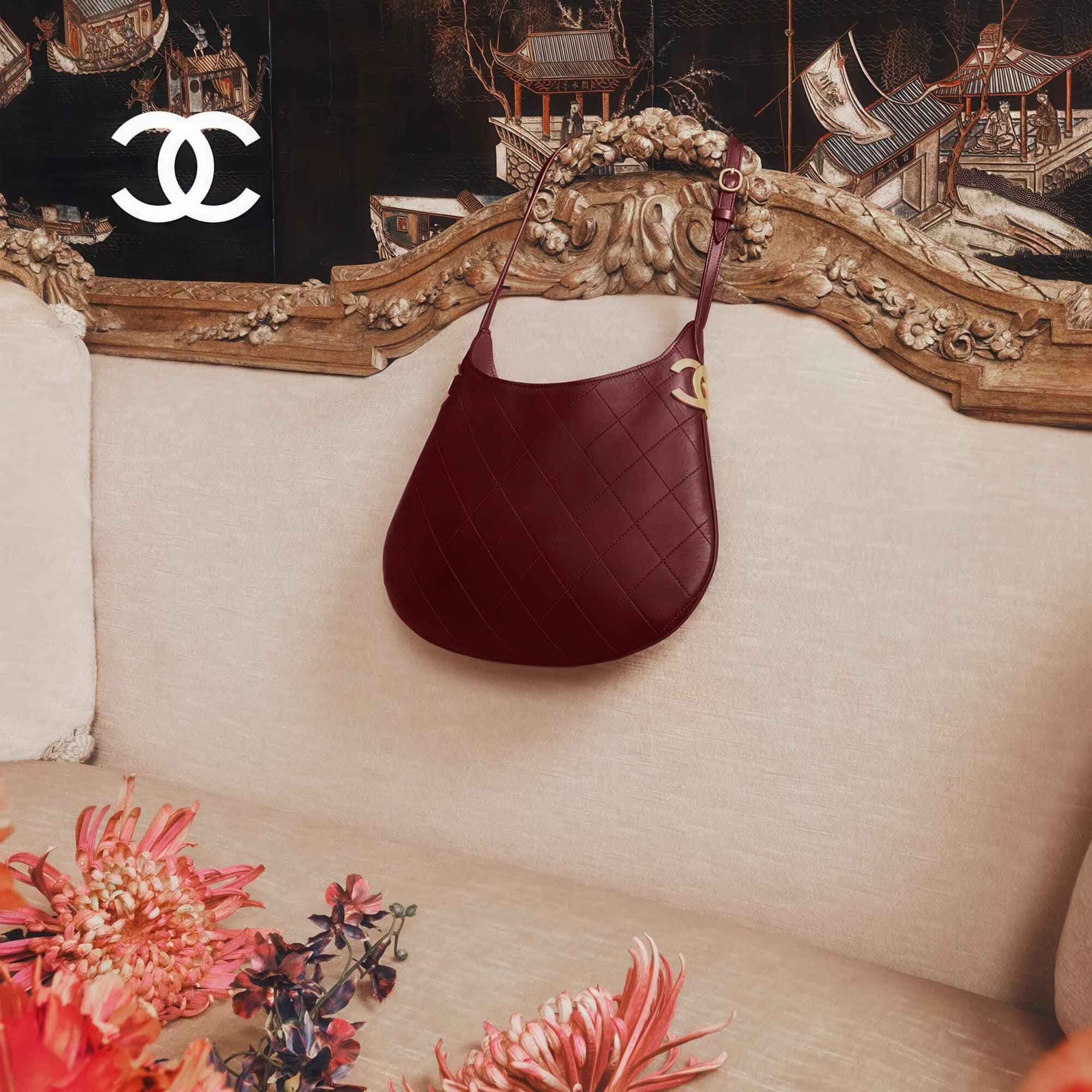 Burgundy quilted leather handbag hung against an ornate vintage sofa with floral arrangements and a black lacquer screen backdrop
