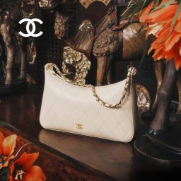 White quilted Chanel handbag with gold chain strap resting on a dark wooden surface with decorative figurines.