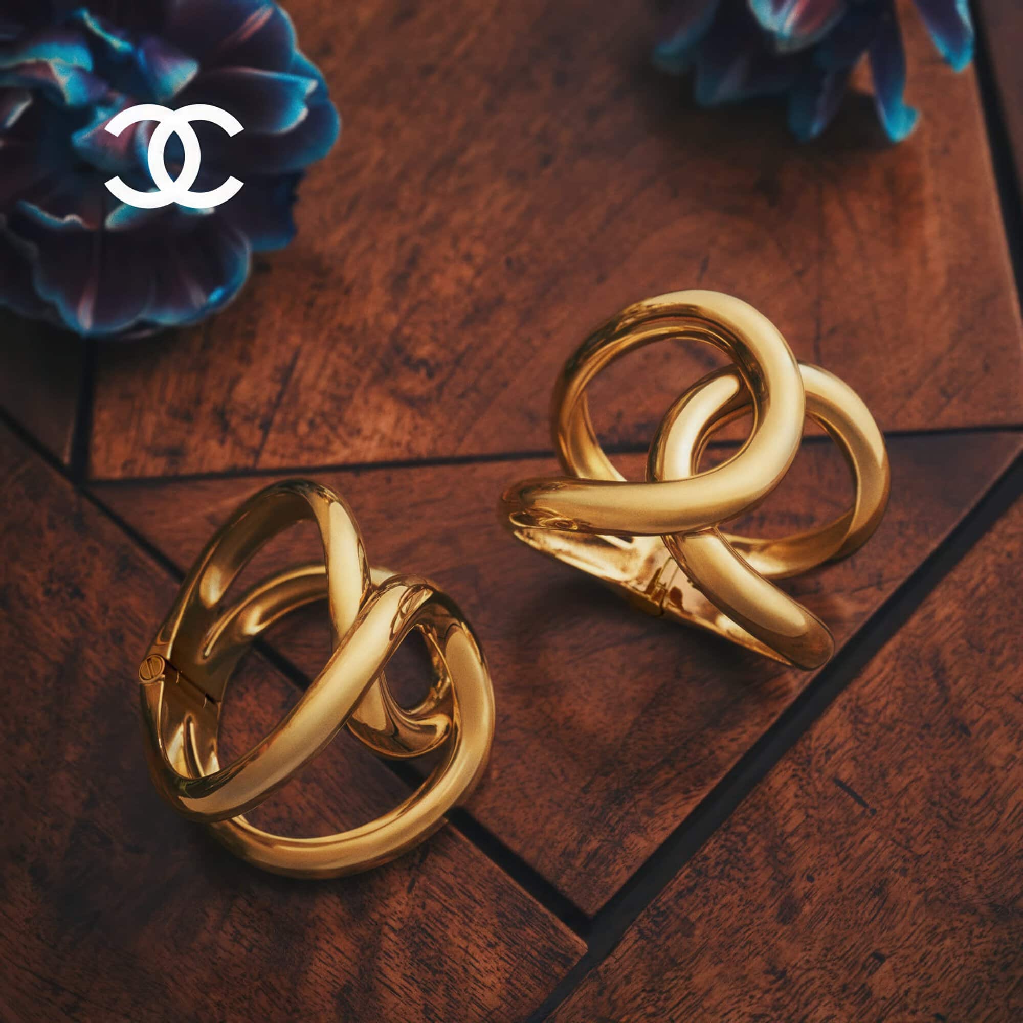 Two intertwined gold hoop bracelets resting on a dark wooden surface next to blue flowers.