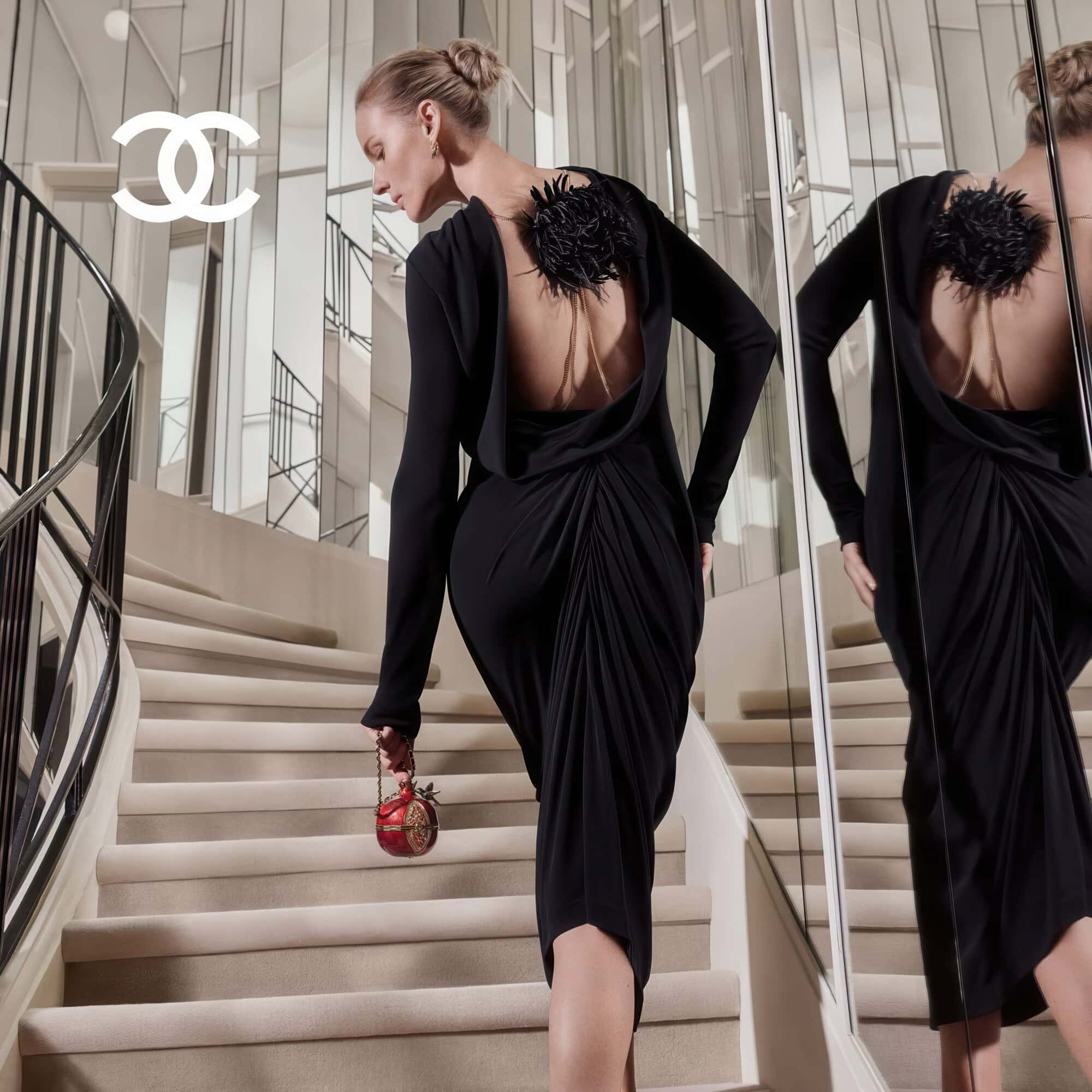 Model in a black cowl-back dress walking up mirrored stairs carrying a red novelty bag for Chanel.