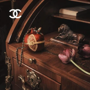 Close-up of a decorative pomegranate-shaped Chanel minaudiere bag on a polished wooden desk with a bronze lion sculpture and a pink tulip.