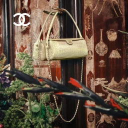 Chanel handbag hanging from a hook against an ornate patterned wallpaper with lush green plants in the foreground.