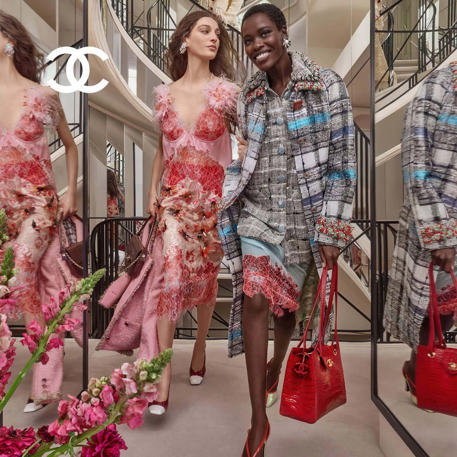 Two models descending a mirrored staircase inside a grand interior, wearing Chanel designer outfits and carrying handbags.