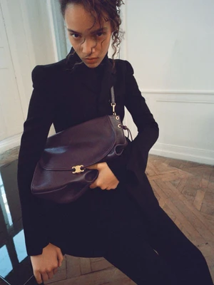 Model in black clothing holding a large dark purple leather shoulder bag in an interior setting