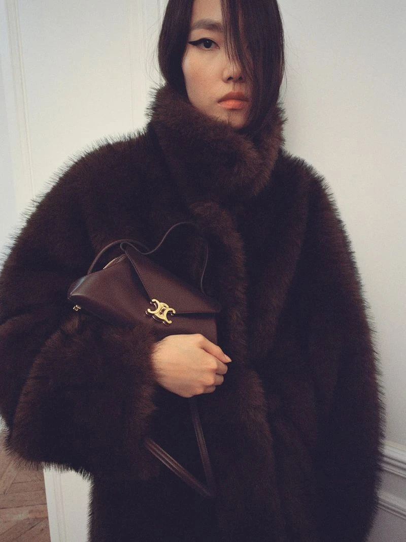 Model wearing a dark brown faux fur coat holding a matching leather handbag with gold clasp for Celine