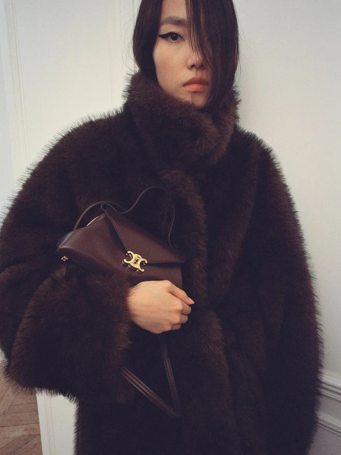 Model wearing a dark brown faux fur coat holding a matching leather handbag with gold clasp for Celine