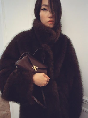 Model wearing a dark brown faux fur coat holding a matching leather handbag with gold clasp for Celine