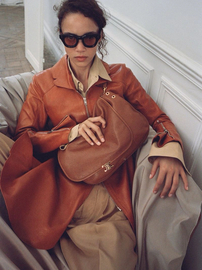 Model wearing dark sunglasses, an orange leather jacket, and holding a brown leather shoulder bag indoors.