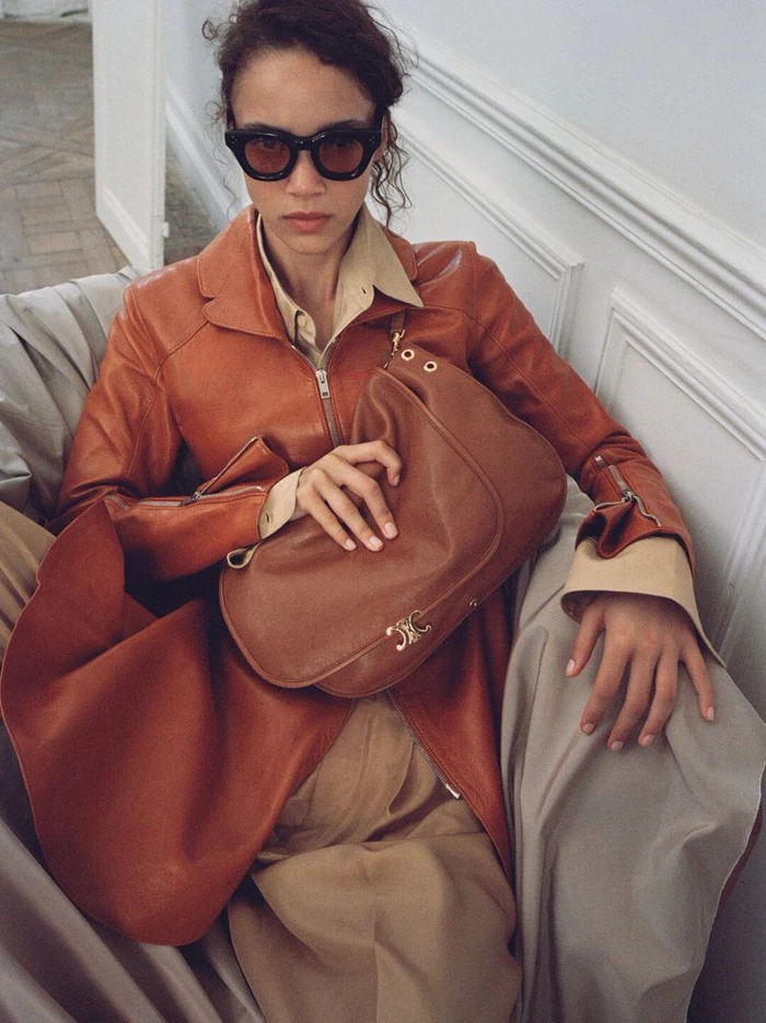 Model wearing dark sunglasses, an orange leather jacket, and holding a brown leather shoulder bag indoors.