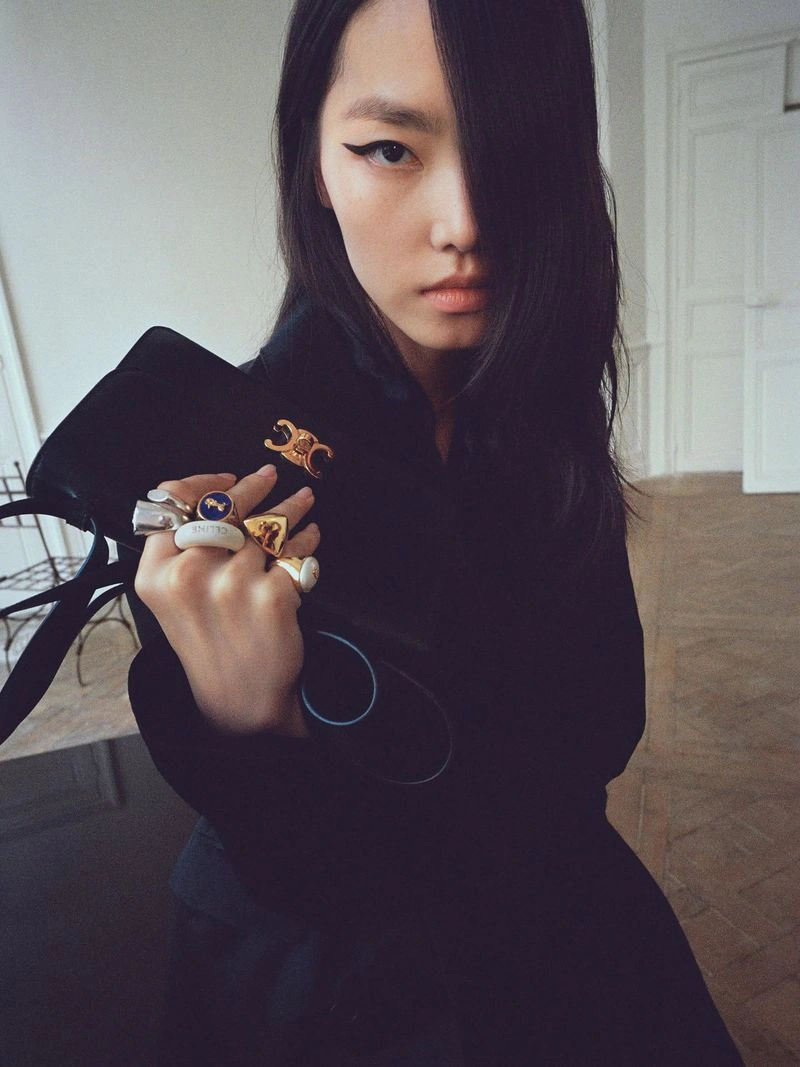 Model with winged eyeliner holding a black handbag adorned with multiple statement rings in an interior setting