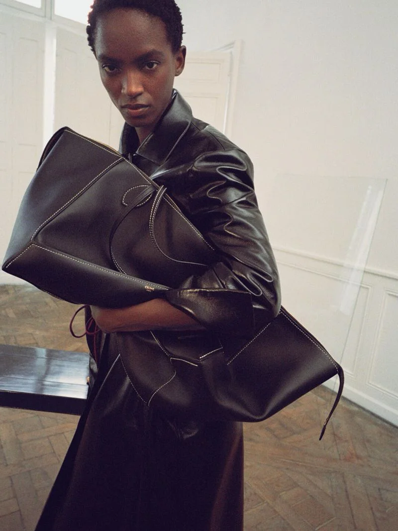 Model wearing a dark leather jacket holding a large black leather Celine bag indoors.
