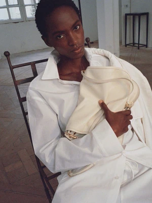 Model seated on a wooden chair holding a white leather shoulder bag for Celine Spring 2027.