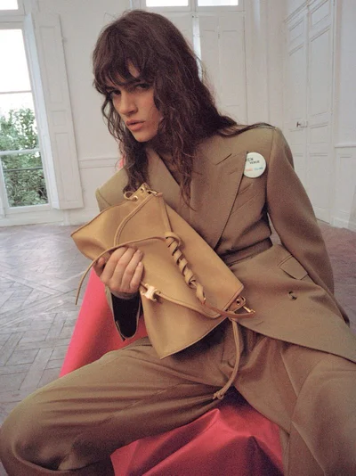 Model wearing a tan suit holding a tan leather bag in an interior space