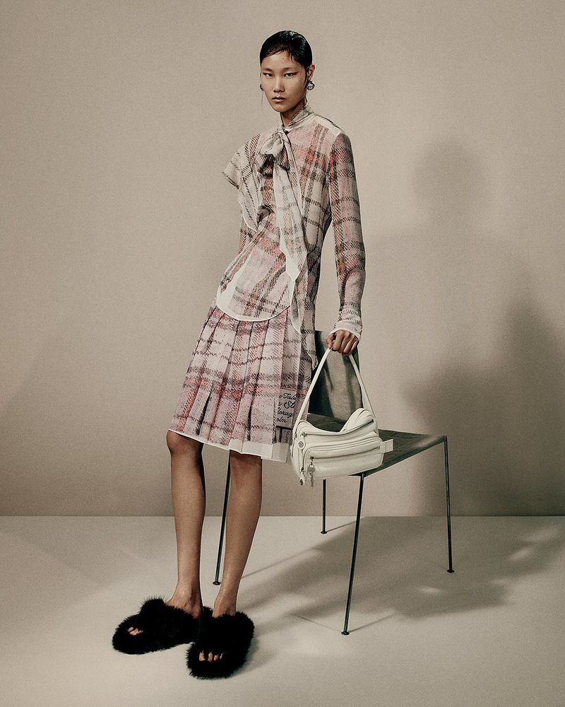 Fashionable Asian woman in plaid top and pleated skirt, holding a white purse, poses in a studio with a modern chair.