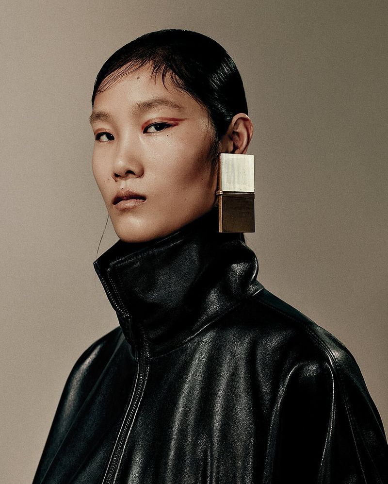 Close-up portrait of an Asian female model with slicked-back hair and red eyeliner, wearing a black leather jacket and large square earrings.