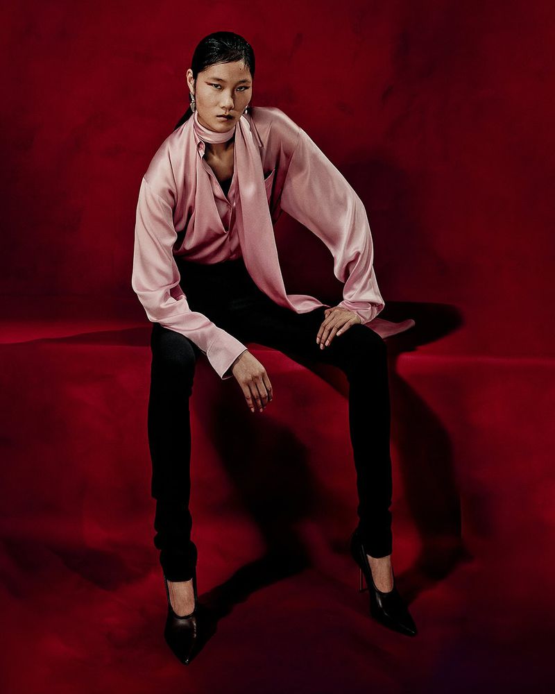 Elegant woman in a pink blouse and black pants sits gracefully against a red backdrop, showcasing a blend of fashion and poise.