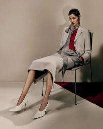 Model Canlan Wang seated in a grey pinstripe suit, red knit top, and white pointed stilettos for Acne Studios F/W 25.