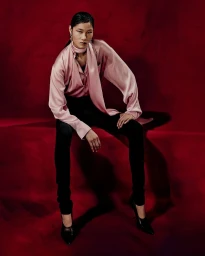 Canlan Wang wears a draped pink satin blouse, black pants, and black heels while seated against a deep red backdrop.