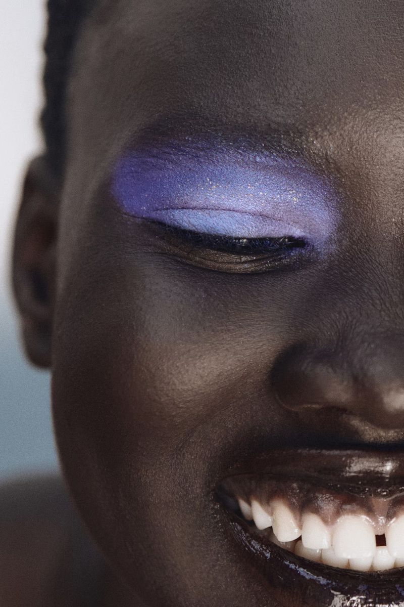 Close-up beauty shot of Nyakong Chan smiling with shimmering purple eyeshadow and glossy dark lips.