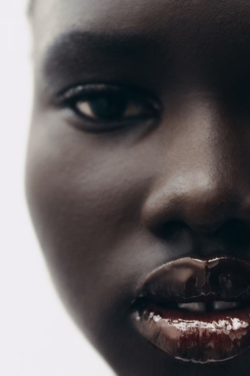 Close-up portrait of an adult woman with dark skin and glossy red lips against a bright background, focusing on beauty and cosmetics.
