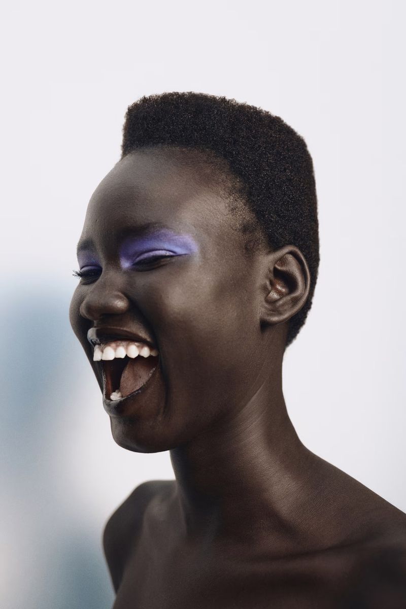 Close-up studio portrait of an African woman smiling with blue eyeshadow and short hair against a blurred, bright background.