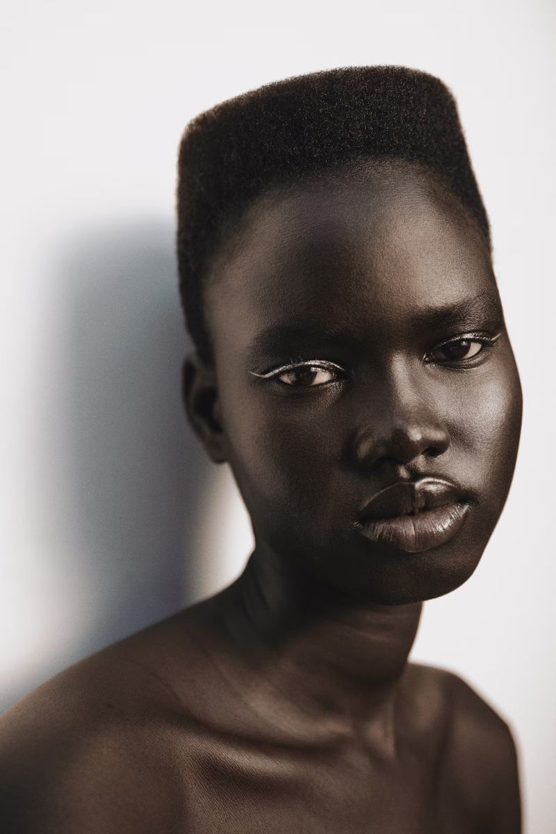 Close-up portrait of a dark-skinned woman with short hair and white eyeliner, against a neutral background with soft lighting, showcasing beauty.