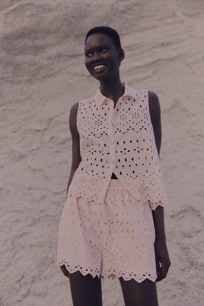 A smiling African woman poses outdoors in a matching pale pink eyelet top and shorts, against a textured background in natural light.