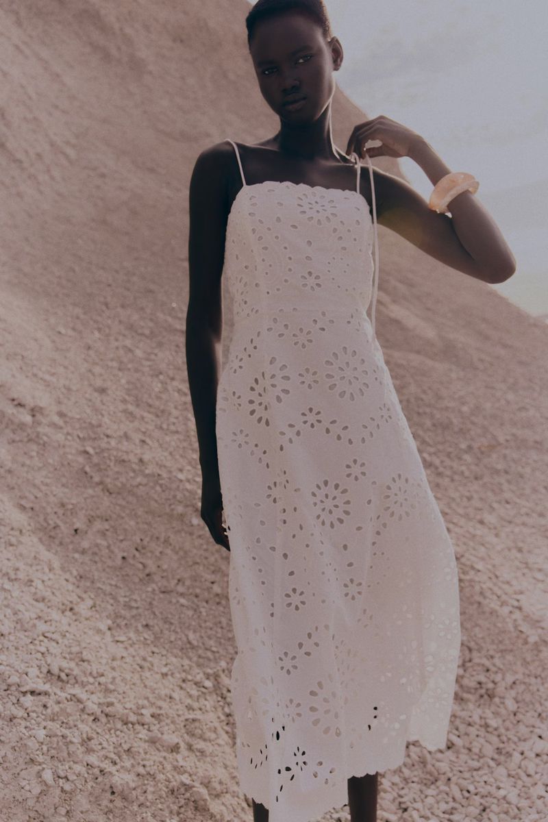 A woman in a white eyelet dress stands in a desert landscape with soft, diffused lighting, showcasing fashion and natural beauty.