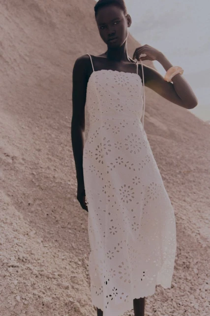 A woman in a white eyelet dress stands in a desert landscape with soft, diffused lighting, showcasing fashion and natural beauty.