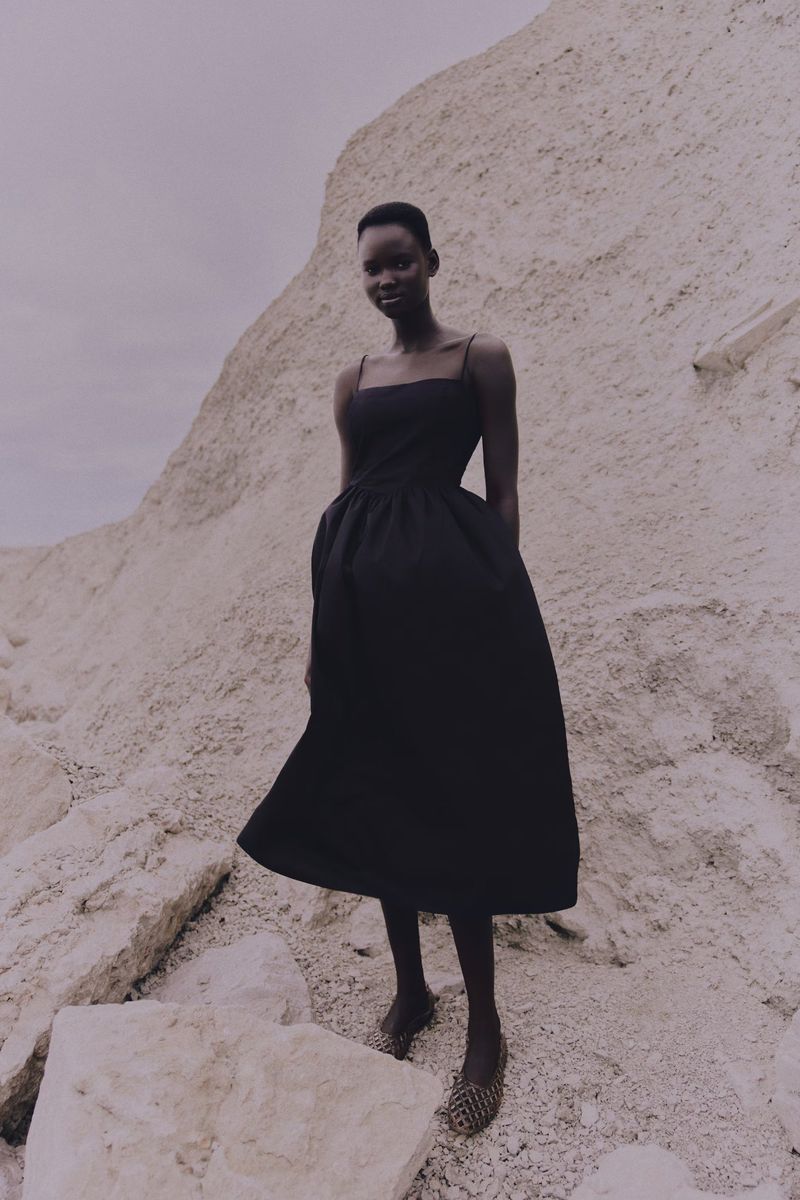 A slender Black woman in a black dress poses on a rocky, sandy hill. The portrait features a gray sky and muted color palette.