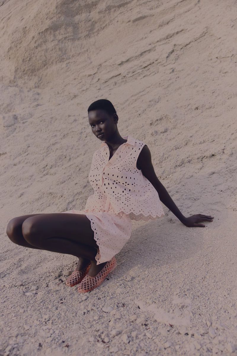 Woman in matching top and shorts sitting in front of a sandy hill. She wears orange see-through flats and poses in natural light.
