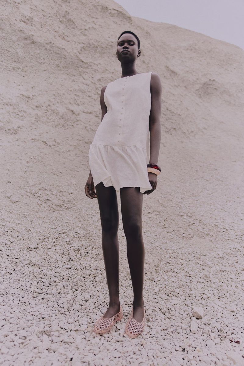 Young adult woman in a sleeveless dress stands in a full shot in a desert environment, with a sandy backdrop. Fashion photography.