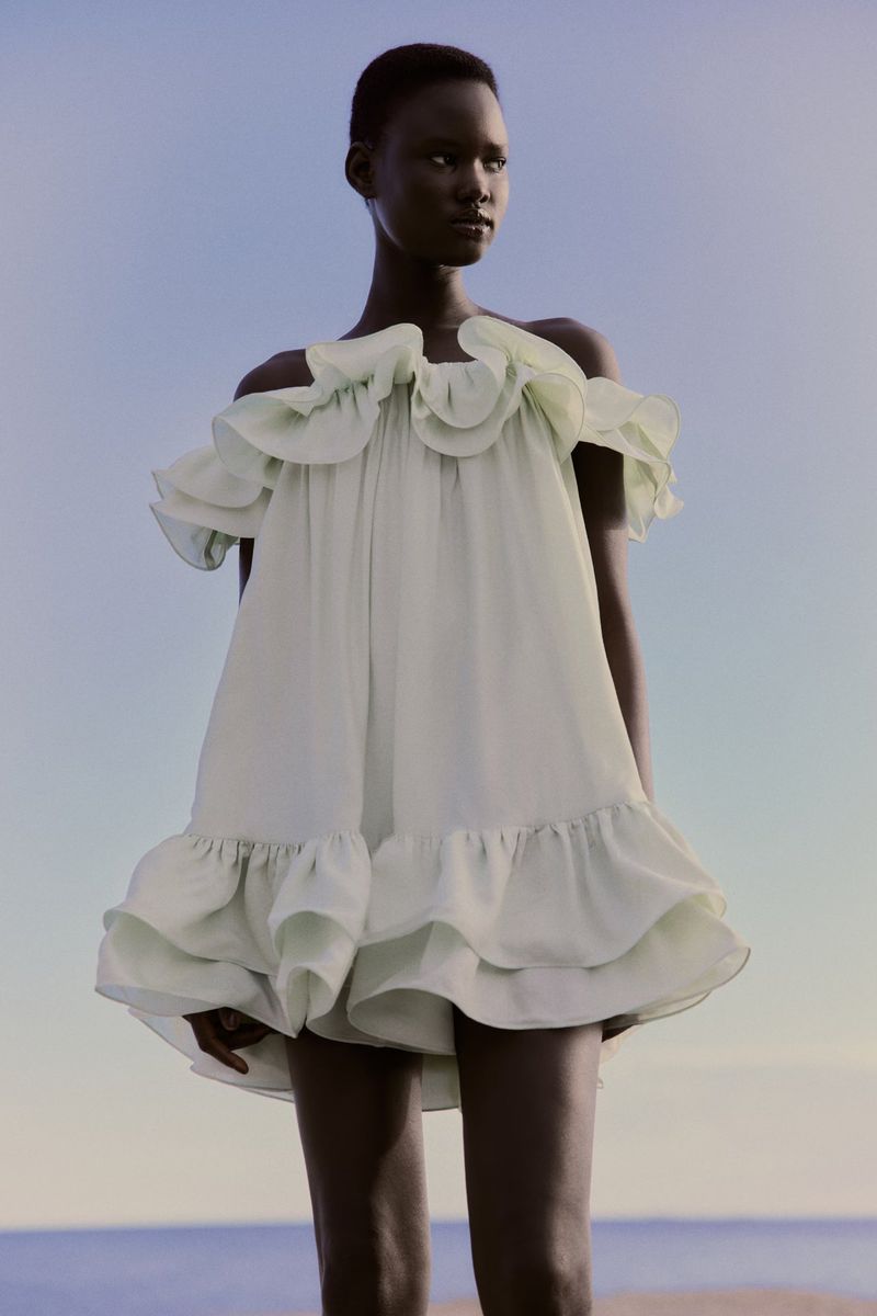 Nyakong Chan wearing a pale green ruffled off-the-shoulder mini dress outdoors against a calm coastal horizon.