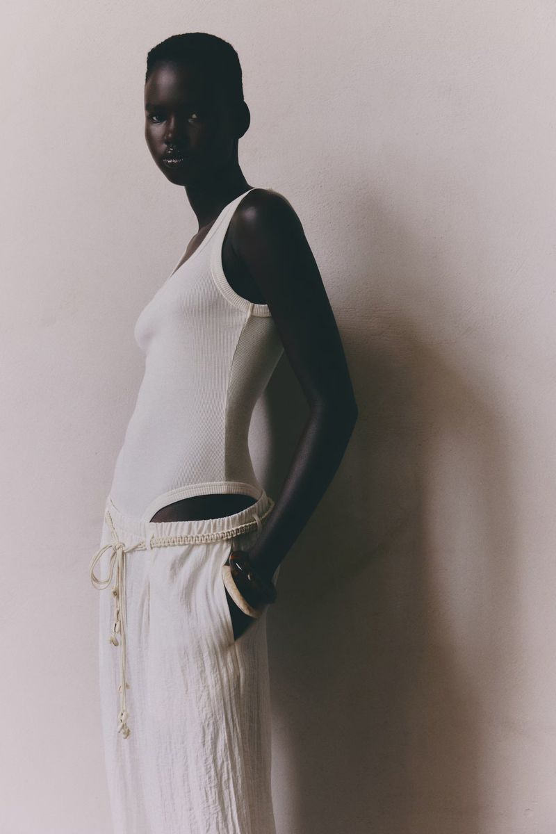 An adult woman poses in a photo studio wearing a white, sleeveless top and pants. The model is lit from the side and slightly above.
