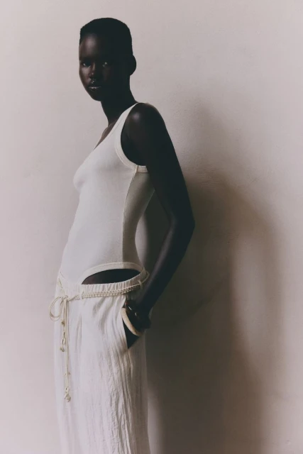 An adult woman poses in a photo studio wearing a white, sleeveless top and pants. The model is lit from the side and slightly above.